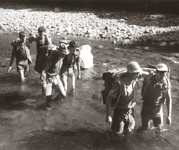 A black and white photo of six trampers lined up in pairs crossing a thigh deep river crossing arms over the top of their packs 