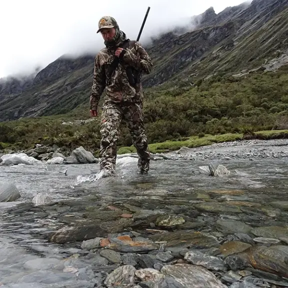 Hunter crossing shallow river with firearm slung on shoulder